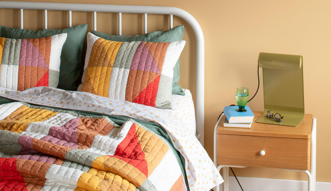 Patterned quilt on bed next to wood side table and perforated green table lamp