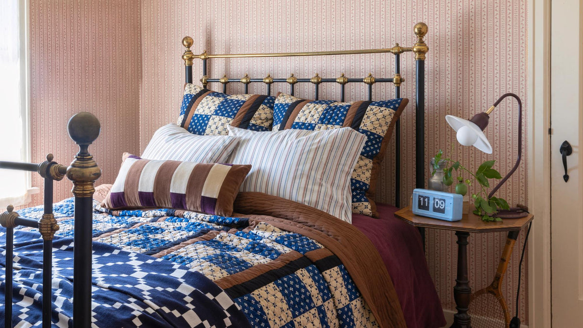 Bedroom with a blue, cream, and brown patchwork quilt on a vintage metal bed against patterned wallpaper