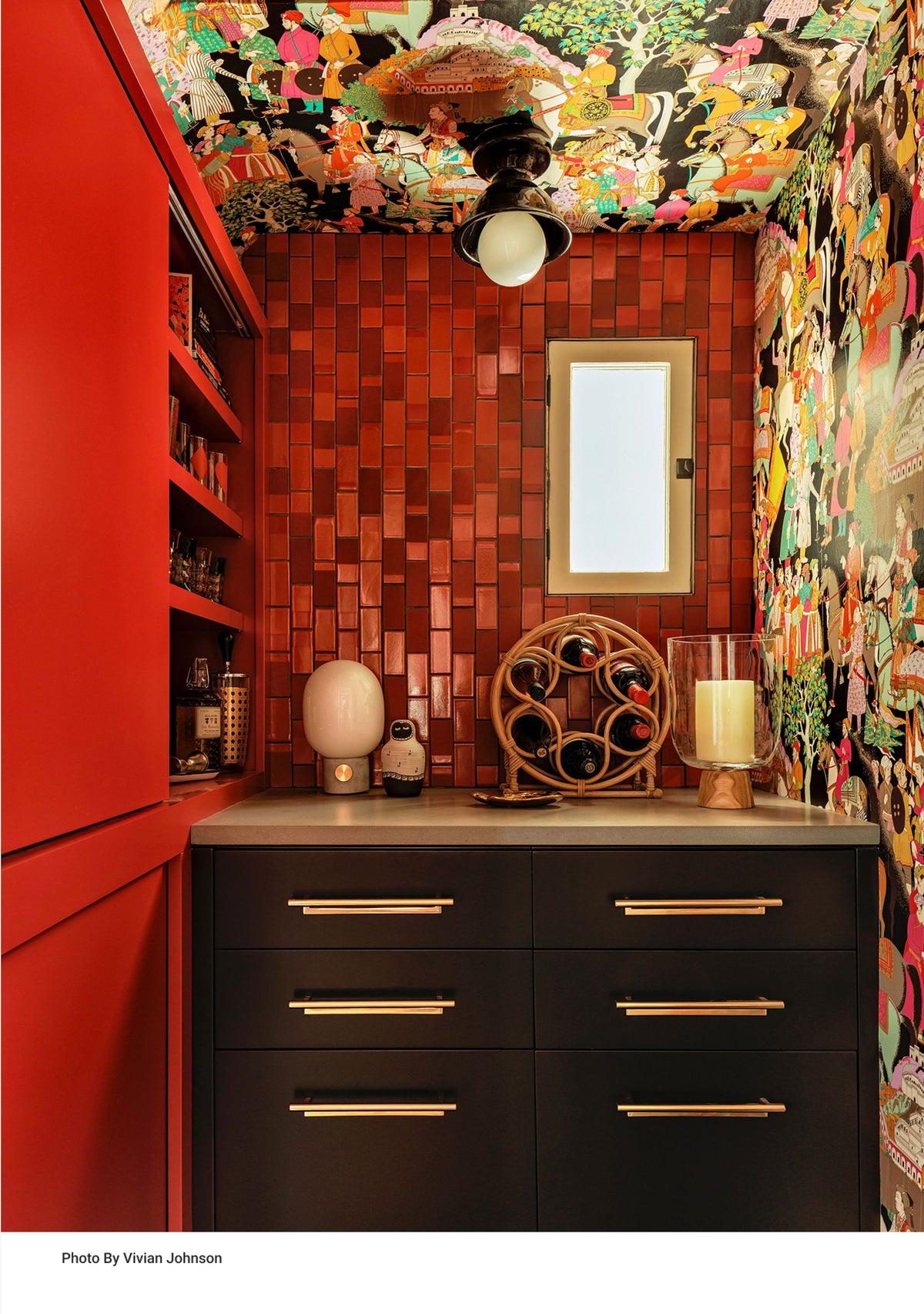 Colorful home bar with red tile, patterned wallpaper, black cabinetry, bottles, and decorative objects