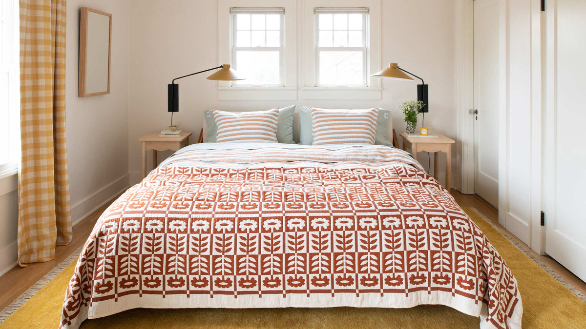Sunlit bedroom with bed styled with rust orange floral quilt, striped pillows, wooden nightstands, and yellow gingham curtains