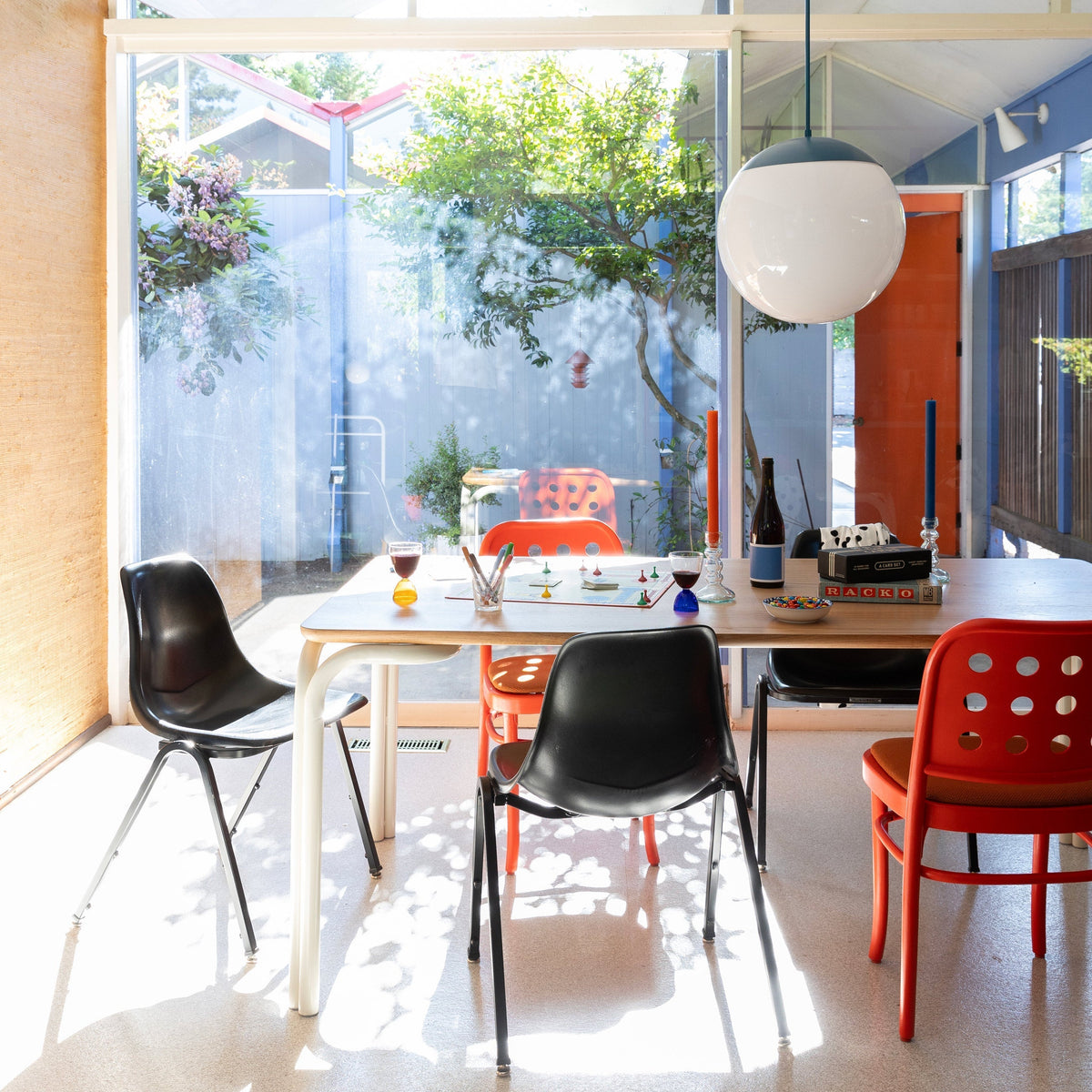 Sunlit dining table with mixed black and red chairs, a board game and wine glasses under a large round pendant light in front of a glass wall