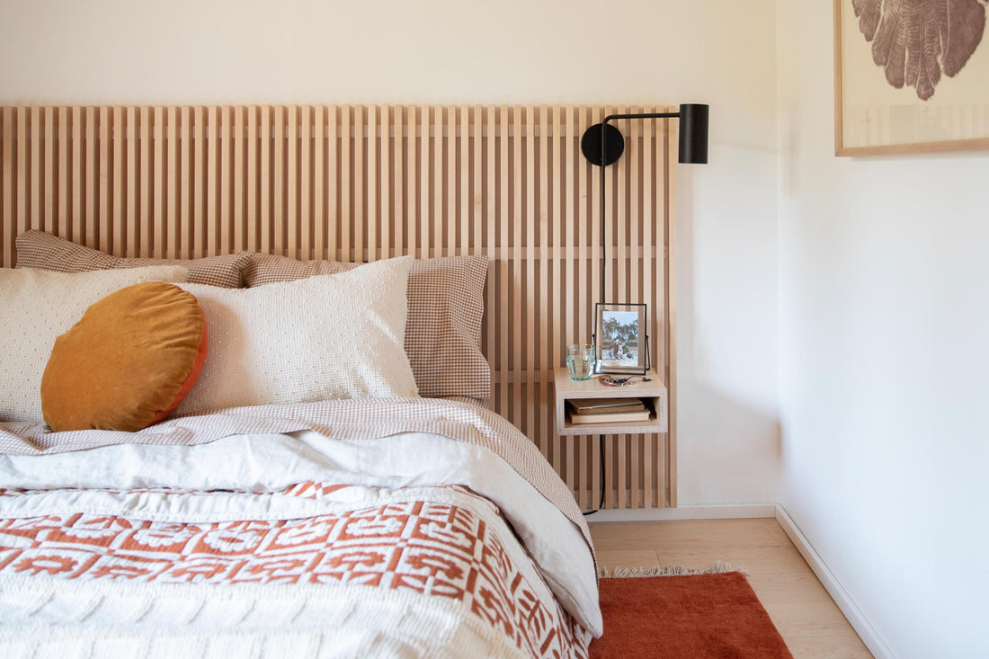 Cozy bedroom with a slatted wood headboard, layered bedding, a mustard round pillow, and a black wall sconce above a floating nightstand