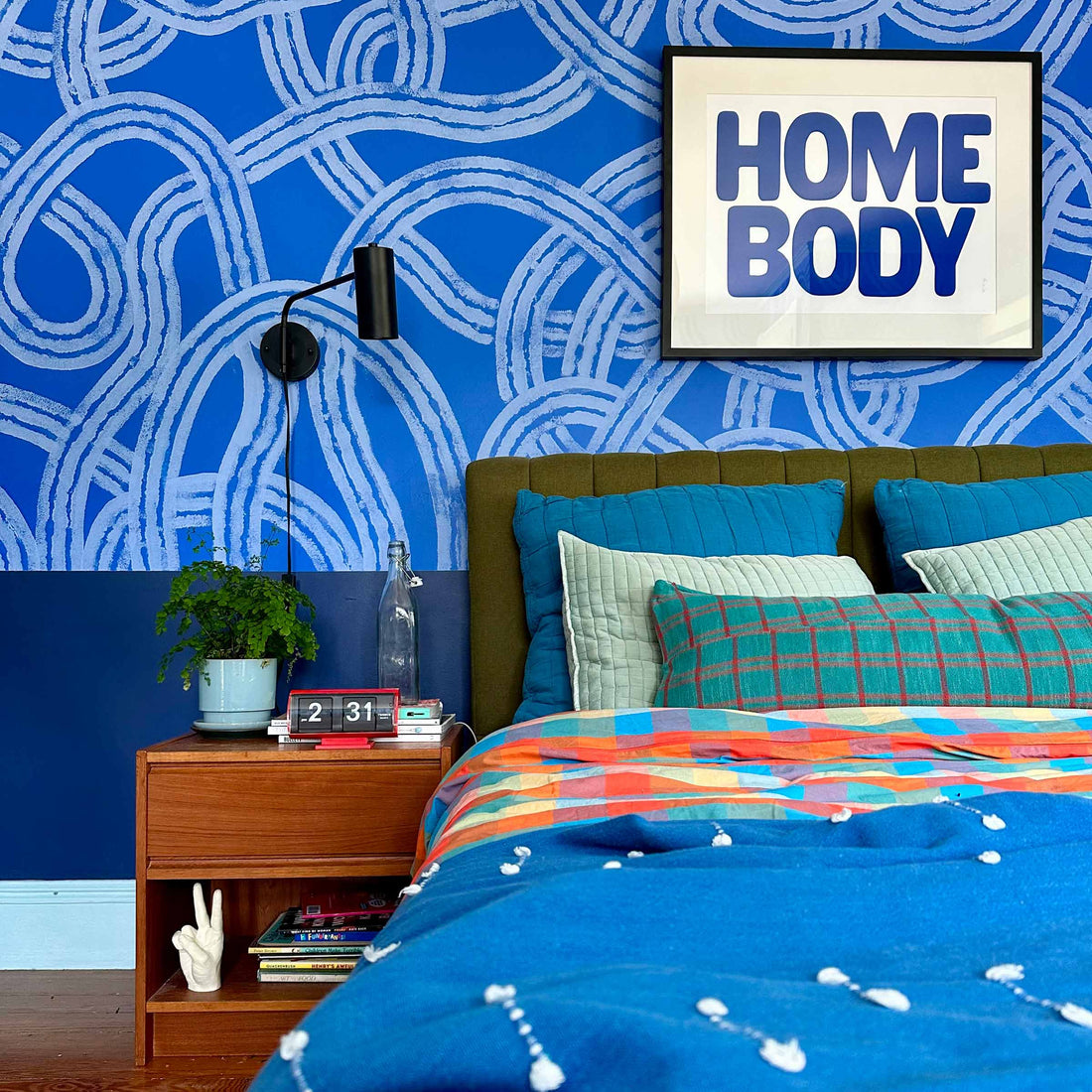 Bedroom with blue looped wallpaper, framed print reading HOME BODY, olive tufted headboard and colorful pillows
