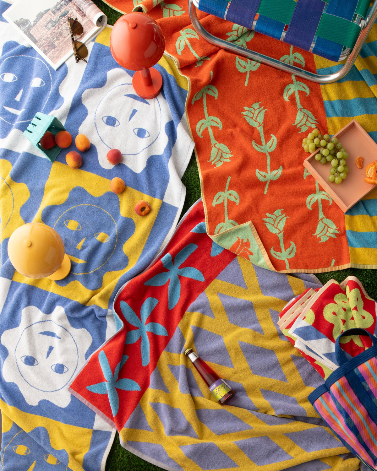 Colorful patterned towels arranged on grass with fruit, lamps, a magazine, and a woven chair