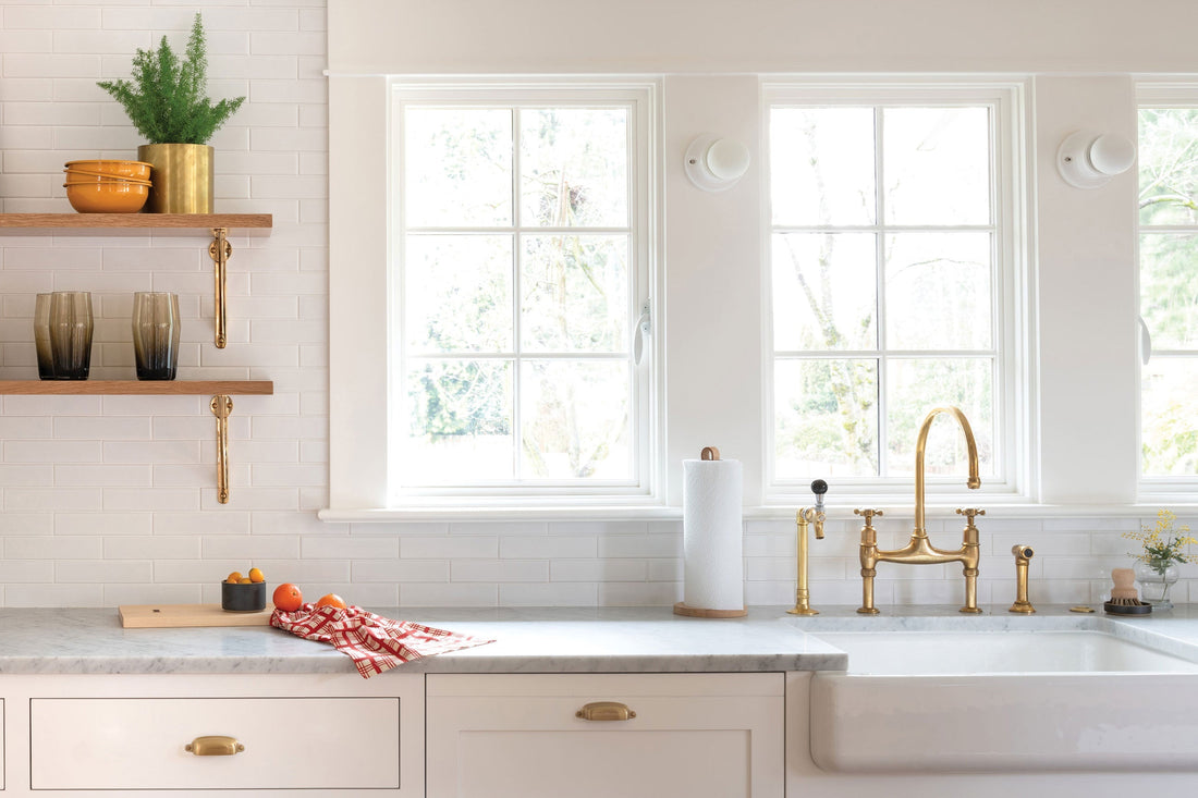 Bright kitchen with a brass bridge faucet over a white farmhouse sink, marble countertop, and open wood shelves with brass brackets