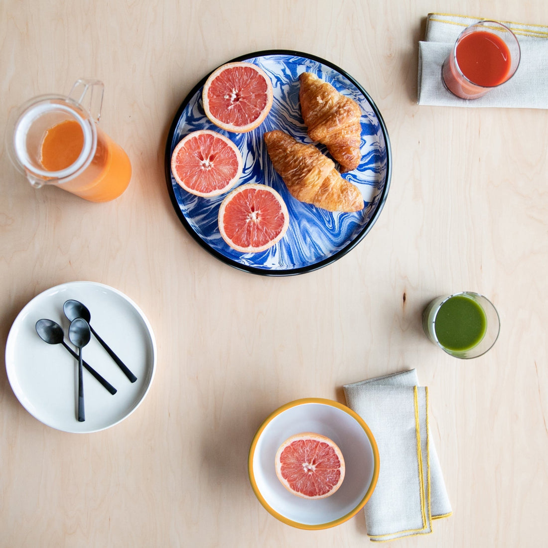 Overhead breakfast flatlay: two croissants and halved grapefruit on a blue marbled plate, juice and bowls on light wood table