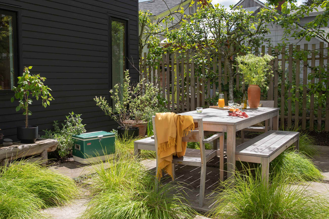 Wooden outdoor dining table with bench and chair draped in a yellow throw set in a fenced backyard with plants and grasses