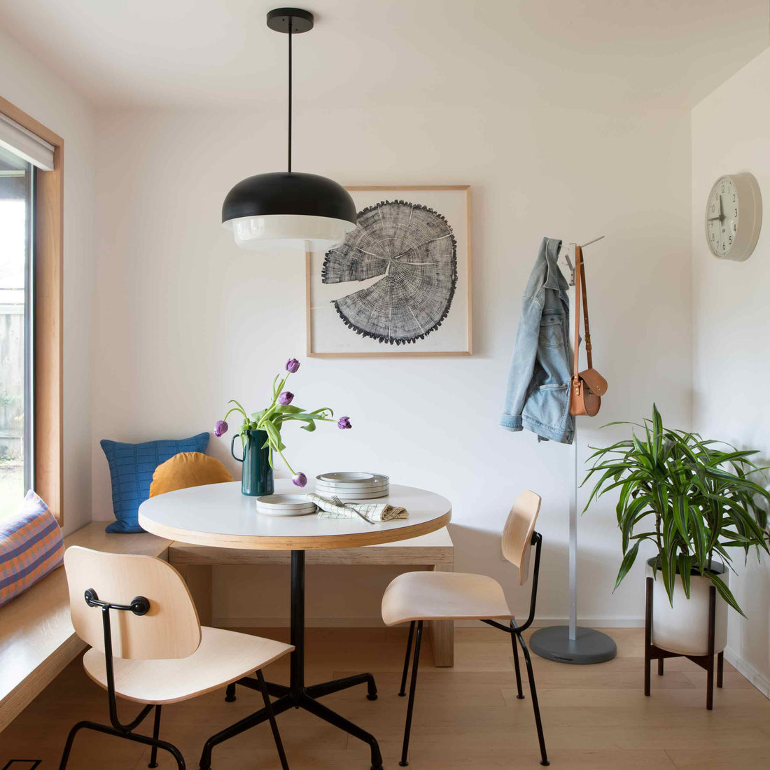 Bright minimalist dining nook with round white table, black dome pendant light, purple tulips in a green pitcher, light wood chairs, coat stand with denim jacket, and potted plant