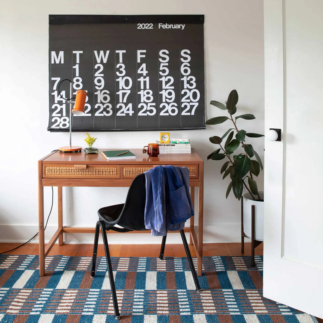 Home office with a wood desk, black chair, wall calendar, lamp, books, plant, and patterned rug