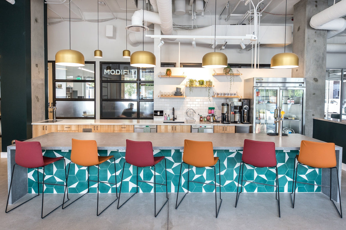Communal kitchen island in a coworking space with teal geometric tile front, six orange and burgundy bar stools, brass pendant lights, and a coffee station in the background
