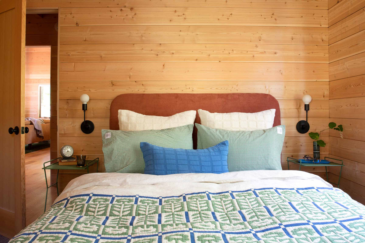 Bed with green and blue leaf quilt, green gingham shams, blue lumbar pillow, and rust upholstered headboard on wood-paneled wall