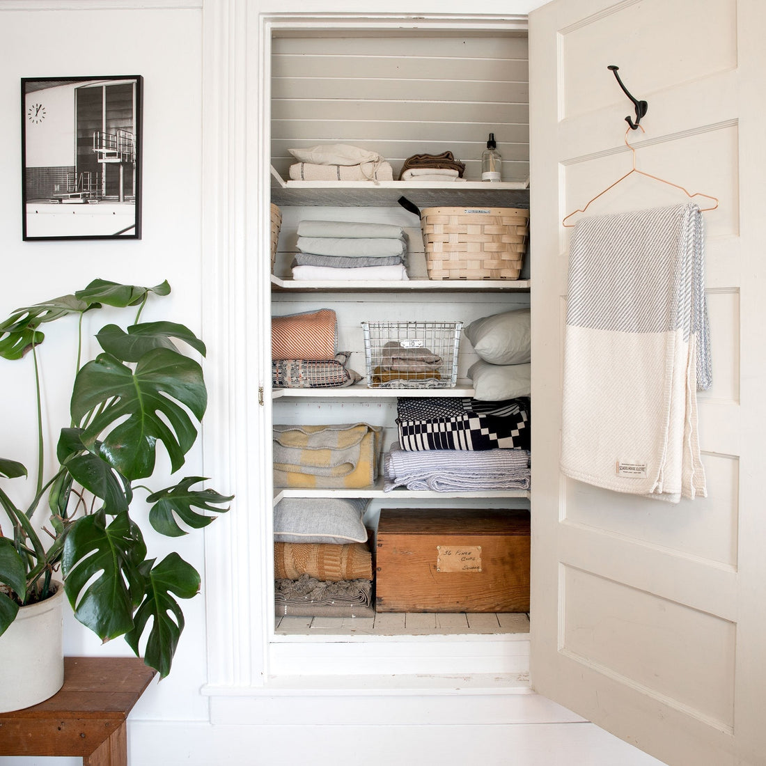 Open linen closet with neatly folded blankets, woven baskets, pillows, and a towel hung on the door in a bright white hallway