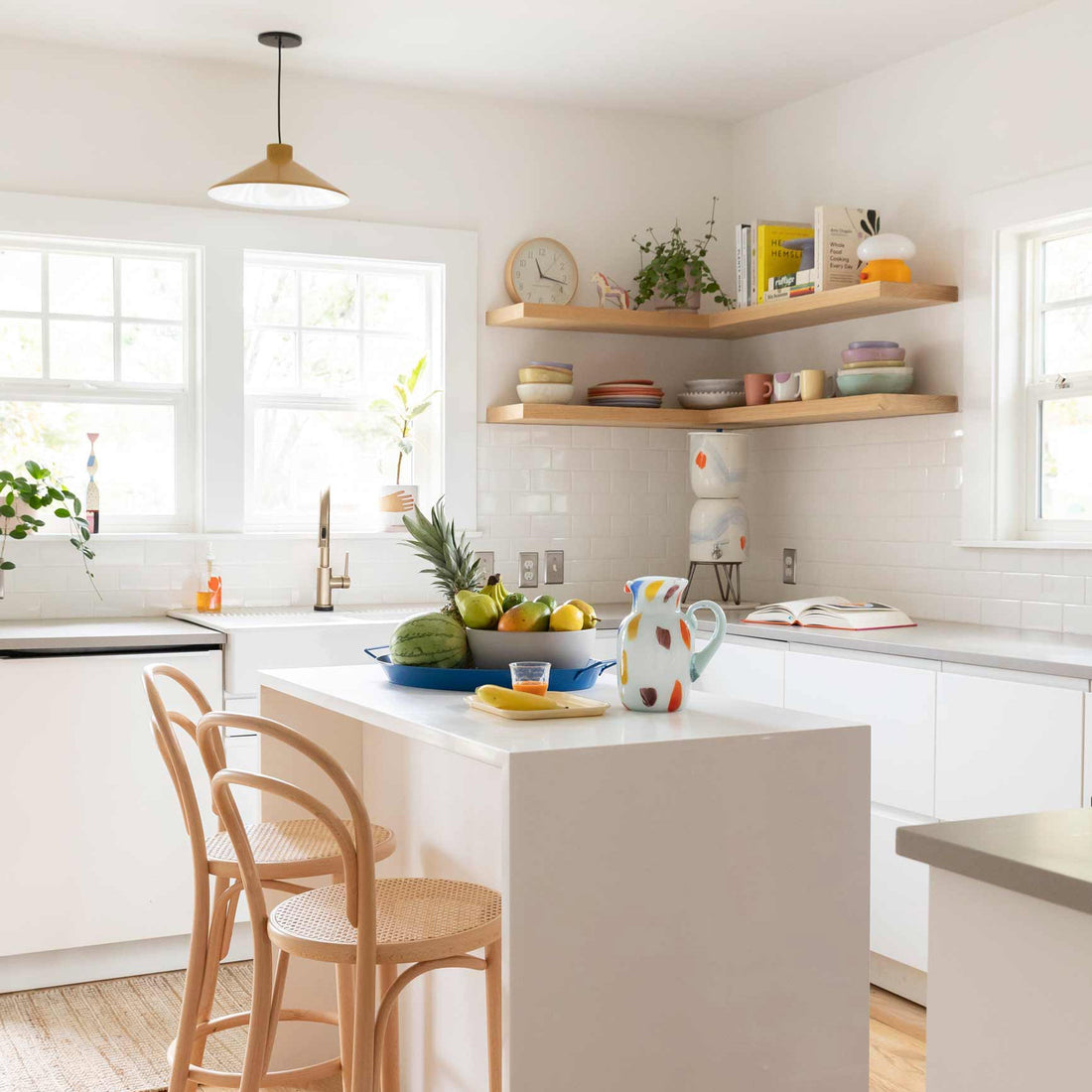 Bright airy kitchen featuring a white island with rattan stools, open wood shelves of colorful dishes, and a painted ceramic pitcher and bowl of fruit on the island