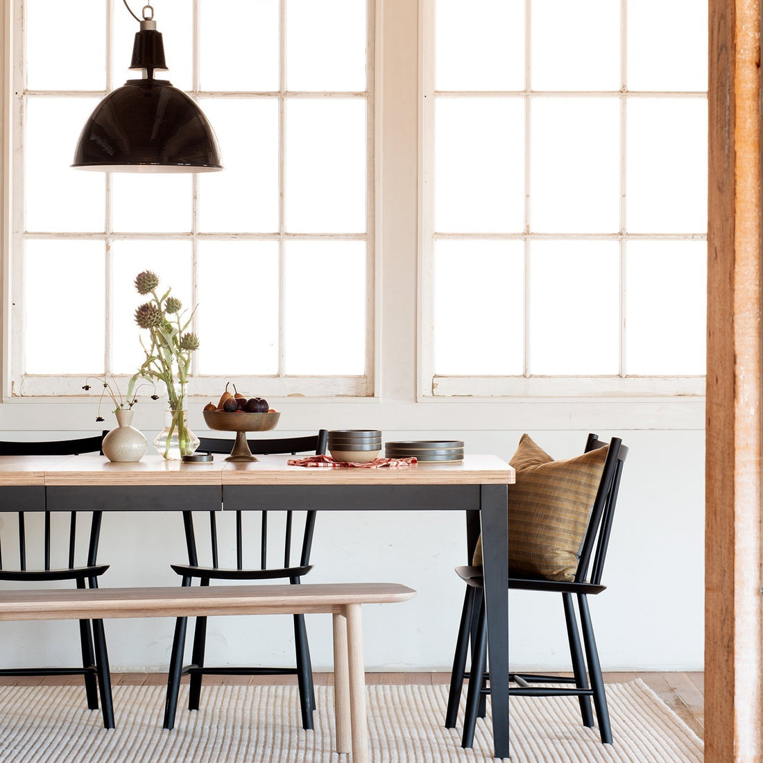 Dining table with black spindle chairs and bench under a glossy black pendant light, styled with vases, a fruit bowl, stacked bowls and a plaid pillow