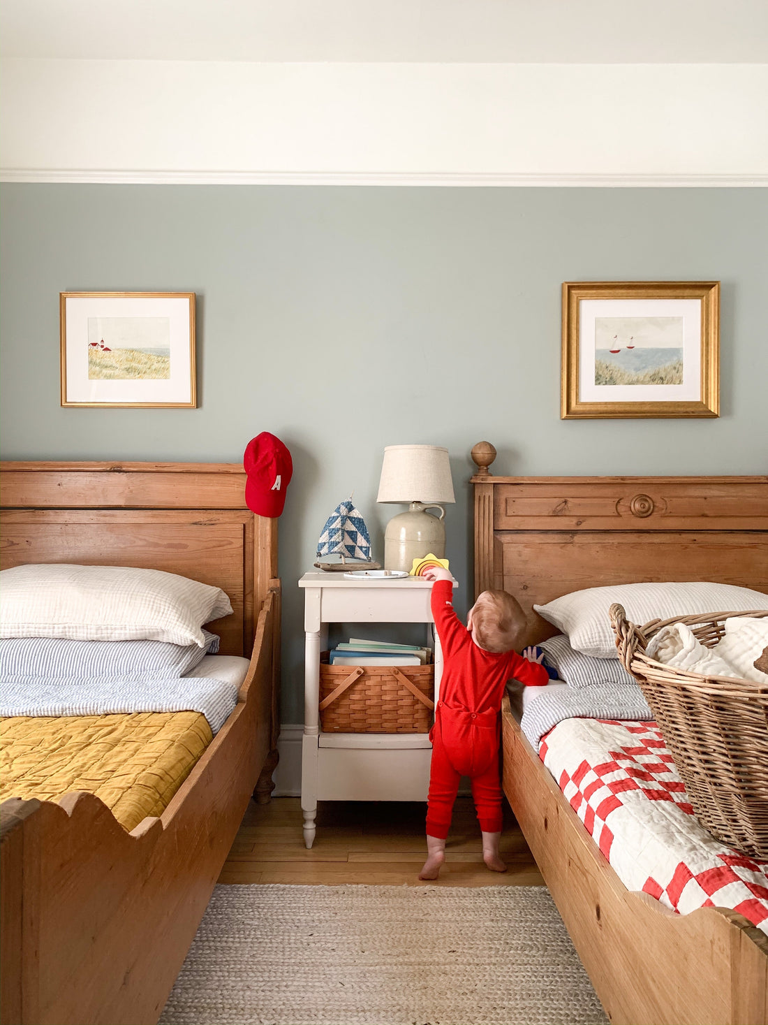 Toddler in red pajamas reaching up to a bedside table between two wooden twin beds in a light green bedroom