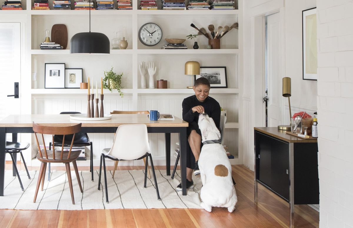 Woman sitting at a dining table smiling and feeding a large white dog in a bright, modern dining room with built-in shelves