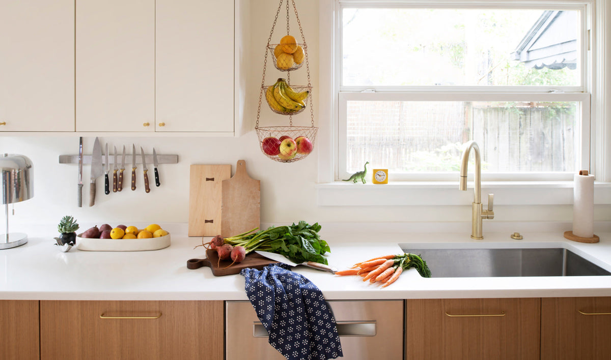 Kitchen countertop with brass faucet and brass cabinet pulls, hanging fruit baskets, cutting boards, and fresh vegetables on a white quartz counter