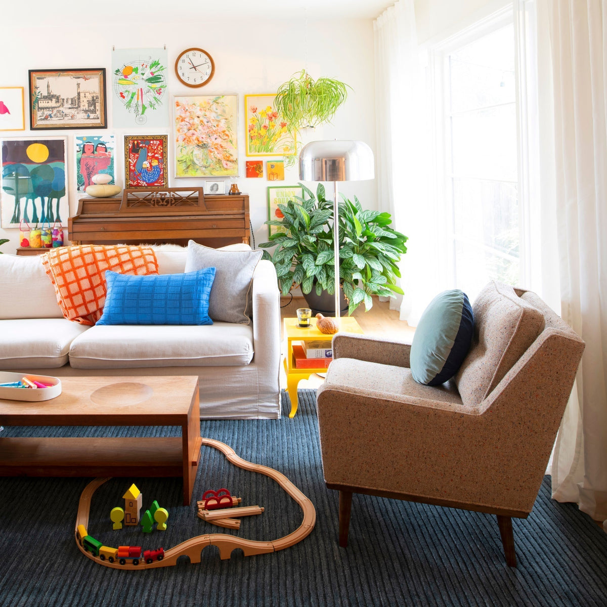 Sunlit living room with white sofa and colorful pillows, armchair, wooden coffee table, toy wooden train on rug, gallery wall