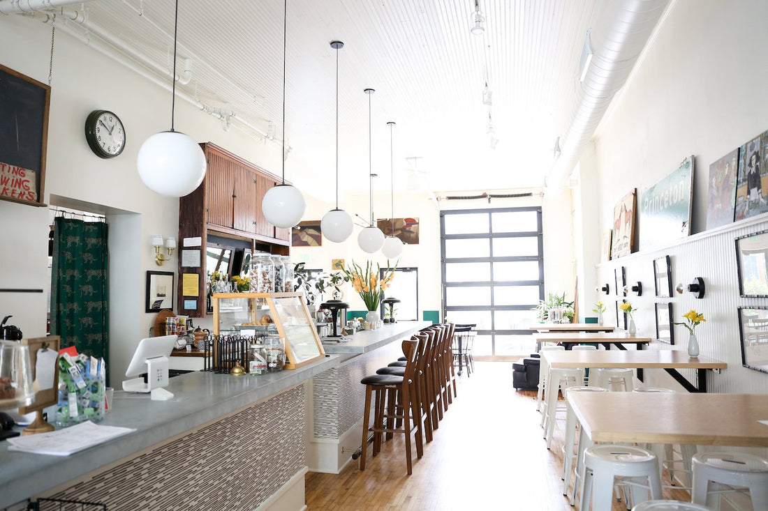 Bright airy cafe interior with long counter, row of wooden stools, globe pendant lights, and large roll-up window