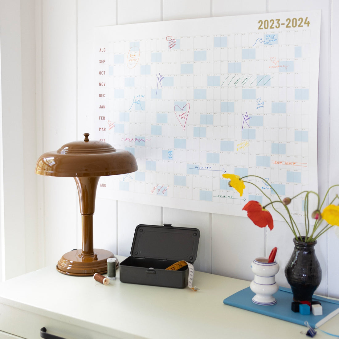 Wall calendar labeled 2023-2024 displayed above a desk with a brown lamp, open black tin, sewing thread, and a vase of flowers