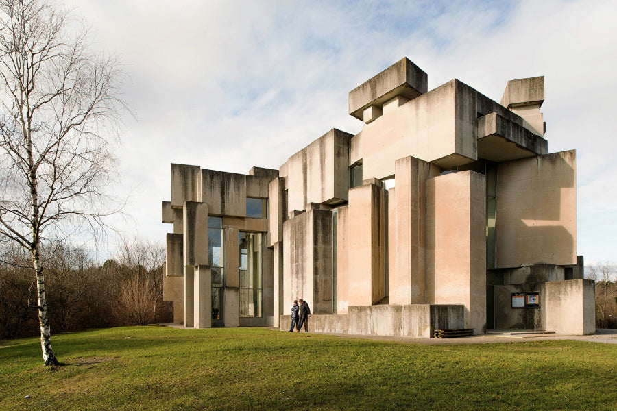 Brutalist concrete building with stacked geometric volumes, two people near the entrance on a grassy lawn under a cloudy sky
