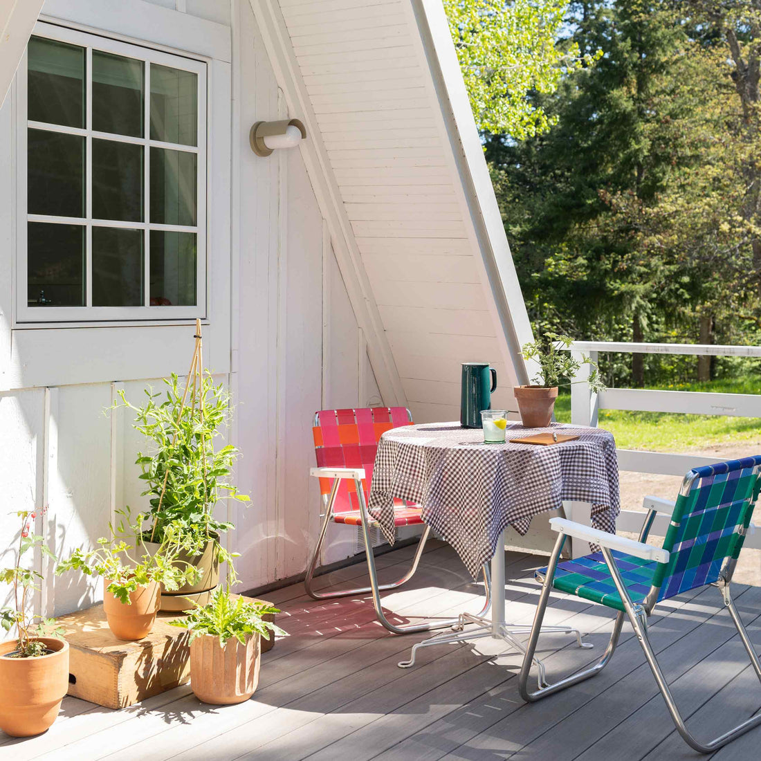 Sunlit porch with round table in checked tablecloth, two colorful woven lawn chairs, potted plants, and forest beyond railing