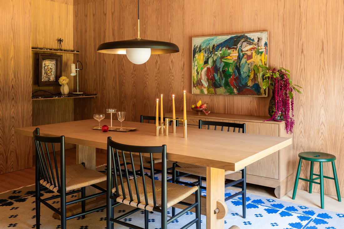 Dining room with oak table and black spindle chairs beneath a dome pendant light, wood-paneled walls, brass candleholders, and a colorful framed painting