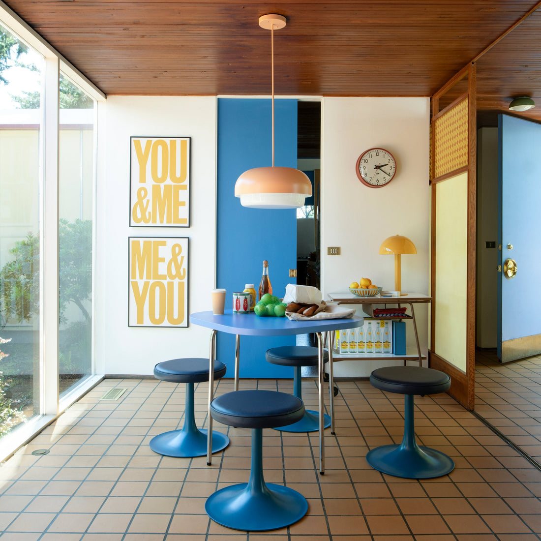 Bright midcentury dining nook with blue round table and pedestal stools, peach pendant light, framed yellow prints reading ‘YOU & ME’ and ‘ME & YOU’, and large floor-to-ceiling windows