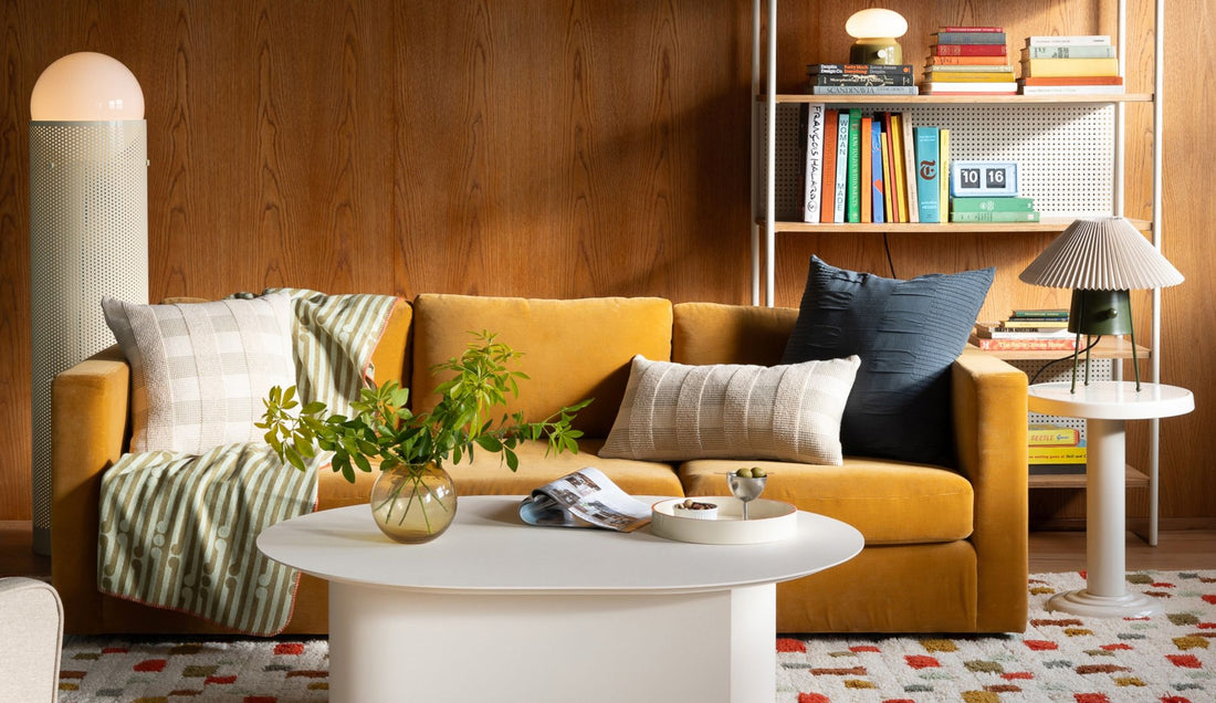 Living room featuring a mustard sofa, white oval coffee table, bookcase, lamp, and patterned rug