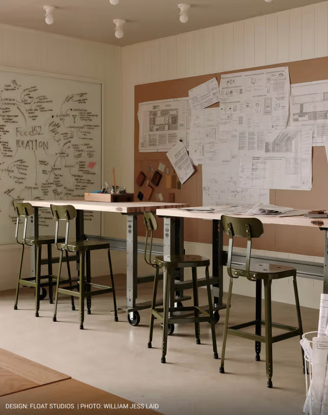 Design studio with two high drafting tables, four olive-green stools, plans on a cork wall and a whiteboard with sketches