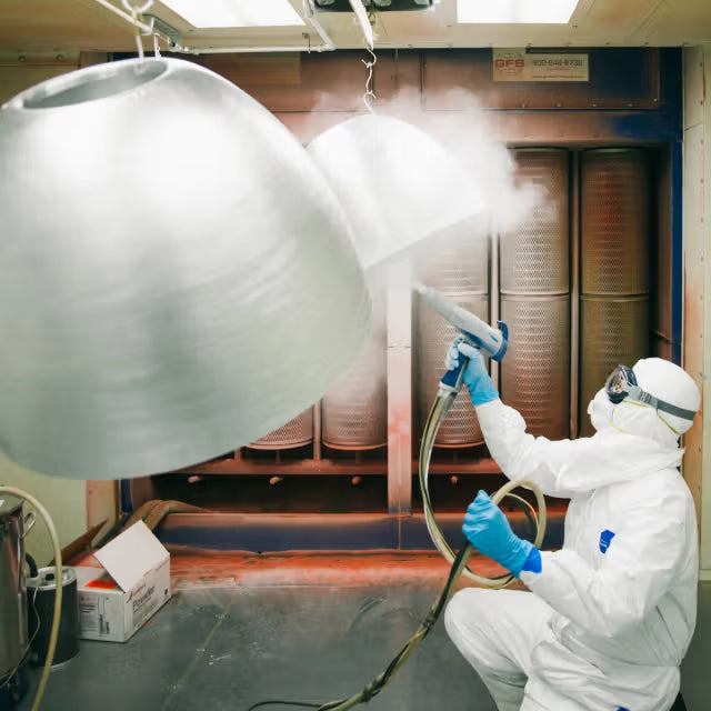 Technician in a white protective suit spray-painting large metal lamp shades inside a paint booth