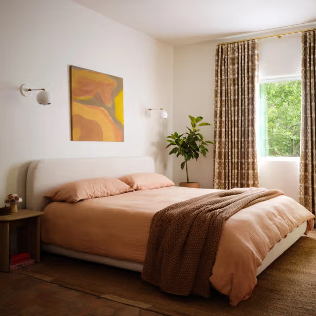 Bed with peach bedding and knitted throw in a sunlit bedroom with wall sconces, patterned curtains, and a potted plant
