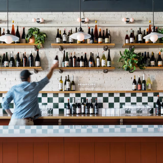 Bartender reaching for a bottle on a wall of wine and liquor shelves behind a tiled bar with pendant lights