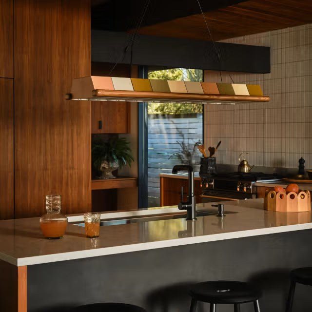 Linear pendant light suspended over a kitchen island with white countertop, black faucet, wood cabinetry, and barstools