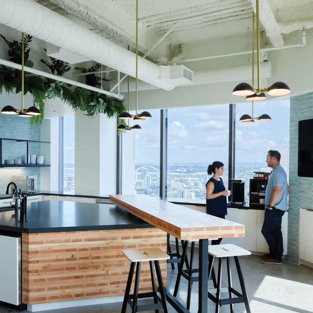 Two people talking by a coffee station in a bright office kitchen with pendant lights, wood-topped island, stools, and skyline view