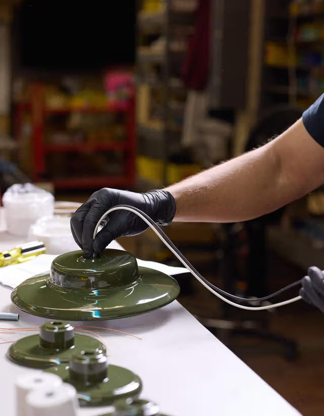 Worker wiring a green metal pendant shade, close-up of gloved hands assembling lighting in a workshop