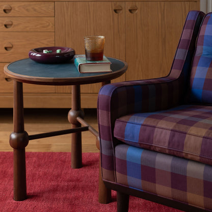 Close up side view of Ann Occasional Table showing dark green leather top and turned walnut legs, styled with a book, amber glass, and a plaid armchair in the background