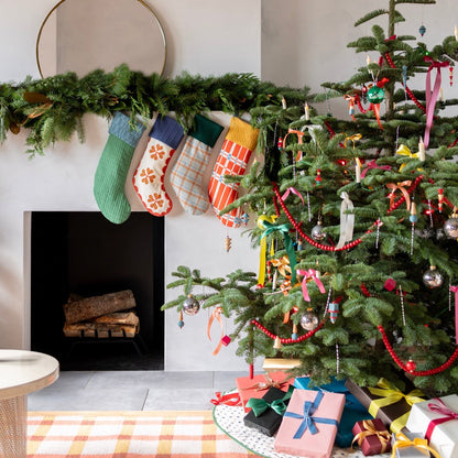 Holiday stockings hang from a greenery-covered mantel beside a decorated Christmas tree and wrapped gifts