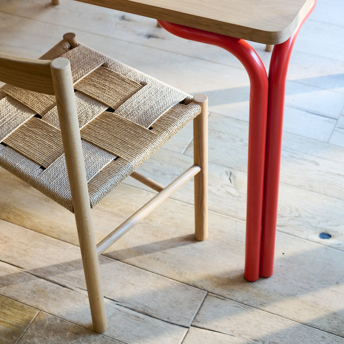 Close-up of Forma Dining Table showcasing its oak veneer top and red powder-coated steel legs beside a woven chair
