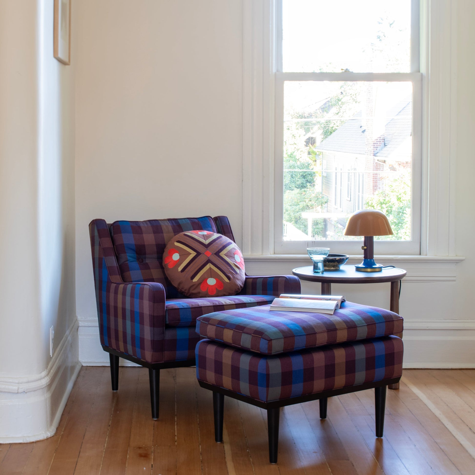 Plaid armchair and ottoman beside a round walnut occasional table topped with a small lamp by a sunlit window