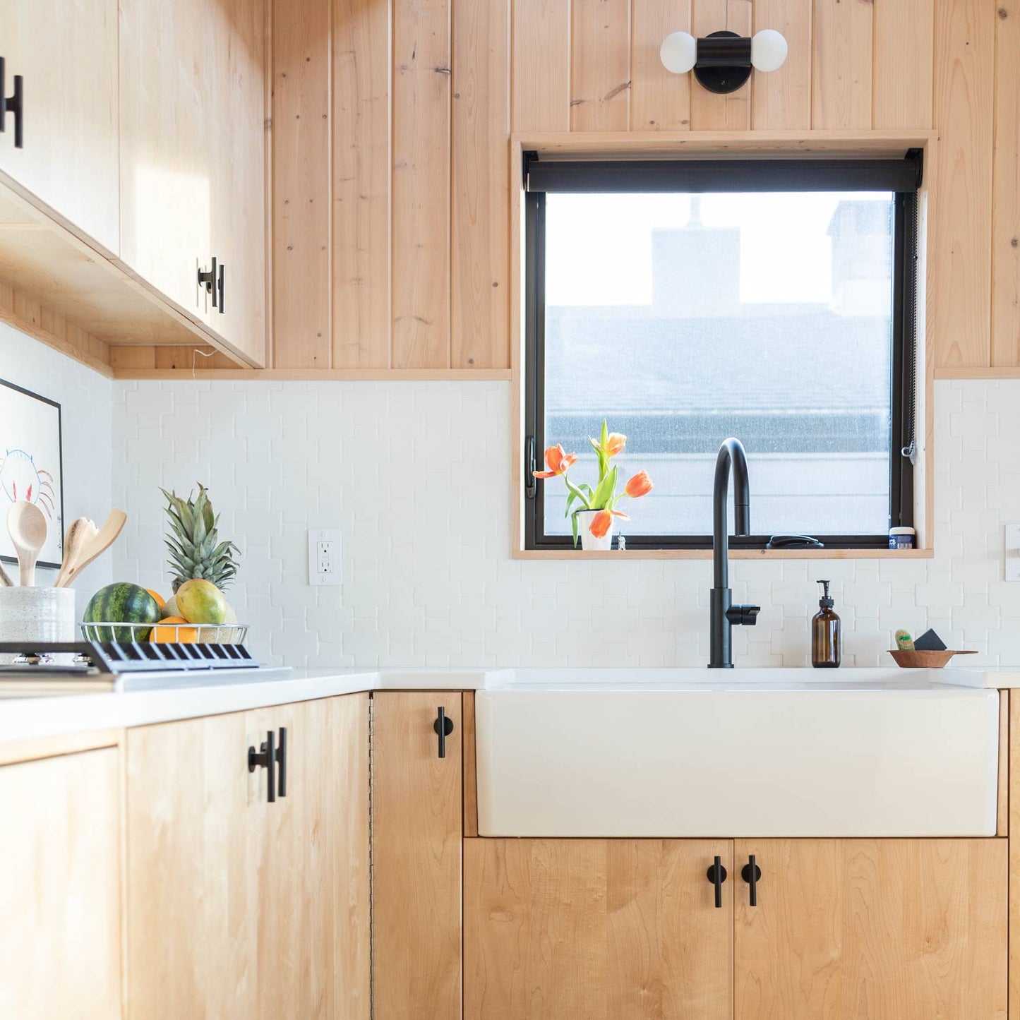 Kitchen sink and countertop with matte black Galley Pull cabinet pulls on light wood cabinets, black gooseneck faucet and orange tulips on the windowsill